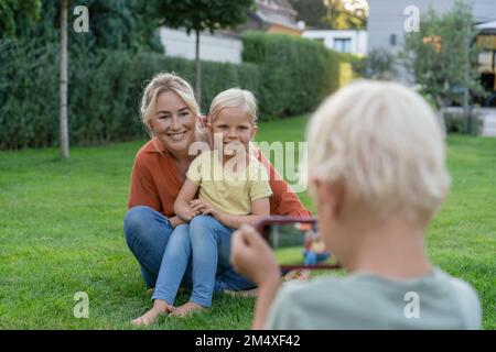 Boy photographing smiling mother and sister through smart phone in garden Stock Photo