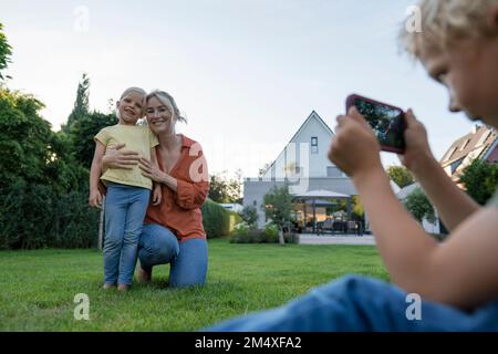 Boy photographing mother and sister through smart phone in garden Stock Photo