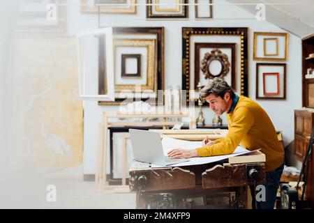 Mature craftsman using laptop and drawing at workbench in workshop Stock Photo