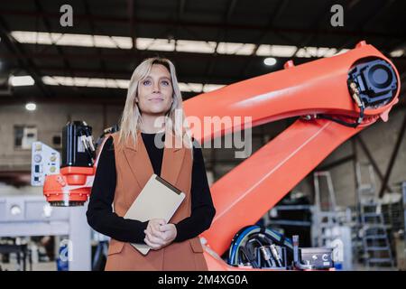 Smiling young engineer with tablet PC standing in front of robotic arm Stock Photo