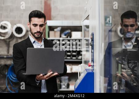 Engineer using laptop standing by machinery at industry Stock Photo