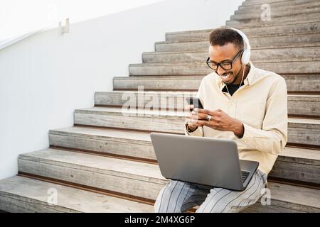 Happy freelancer text messaging through smart phone sitting with laptop on steps Stock Photo