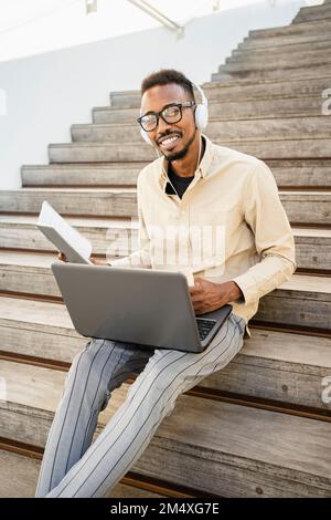 Happy man sitting with laptop wearing headphones on steps Stock Photo