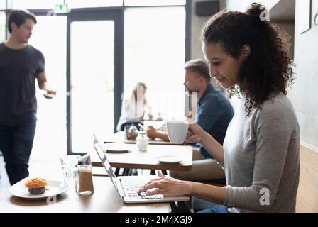 Smiling freelancer working on laptop drinking coffee at cafe Stock Photo