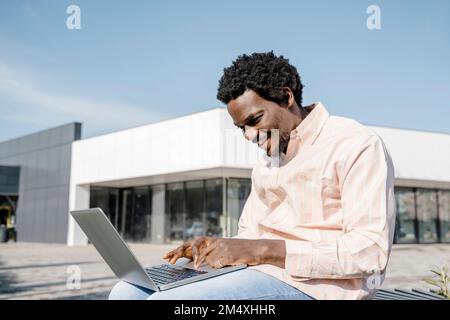 Smiling young businessman using laptop sitting on bench Stock Photo