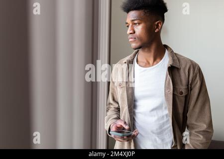 Thoughtful young man with smart phone looking out of window at home Stock Photo