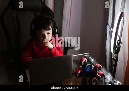 Boy wearing headphones using laptop sitting by mobile phone in illuminated ring light Stock Photo