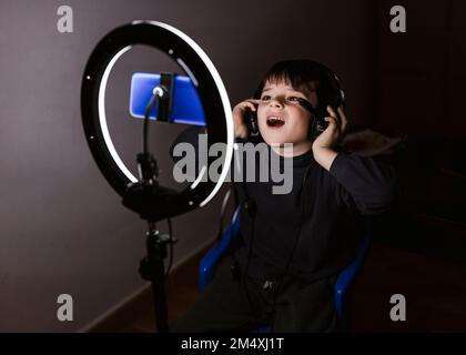 Boy wearing headphones recording video through mobile phone in illuminated ring light Stock Photo