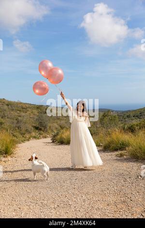 Playful girl wearing white dress holding balloons with dog on pathway Stock Photo