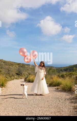 Girl wearing white dress holding balloons playing with dog on pathway Stock Photo