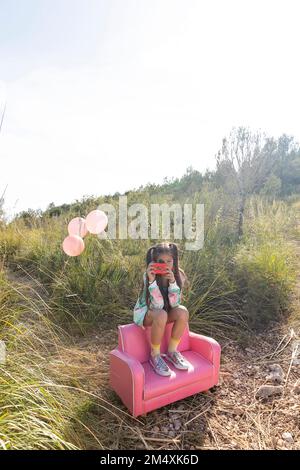 Girl photographing through toy camera sitting on pink armchair Stock Photo