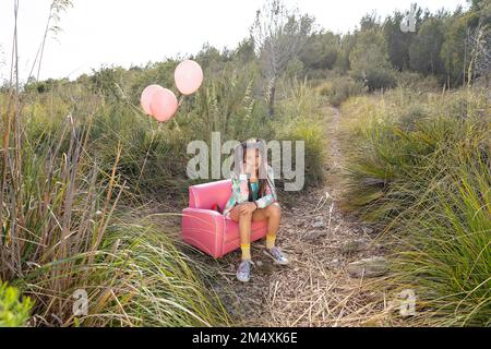 Thoughtful girl sitting on pink armchair in field Stock Photo