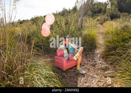 Girl photographing through toy camera sitting on pink armchair in field Stock Photo