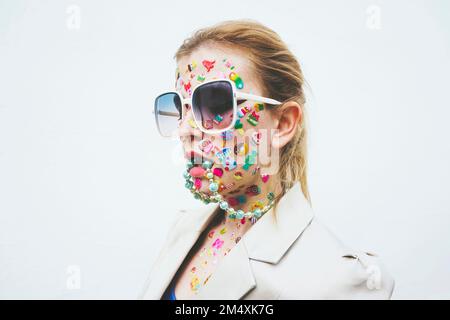 Woman with multi colored stickers on face and necklace in mouth against white background Stock Photo
