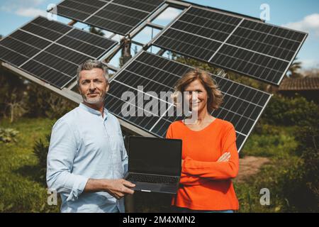 Smiling man and woman with laptop in front of solar panels on sunny day Stock Photo