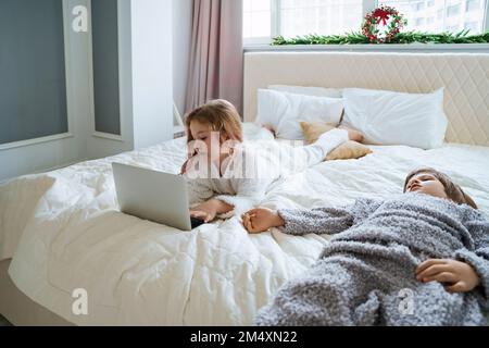 Girl using laptop lying on bed by brother at home Stock Photo