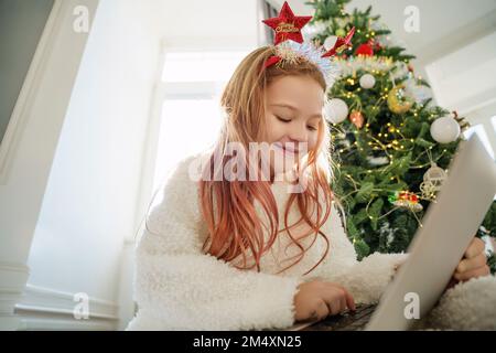 Happy girl using laptop sitting in front of Christmas tree at home Stock Photo