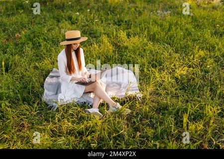 Inspired female author wearing straw hat writing creating on laptop computer sitting on beautiful field of green grass Stock Photo