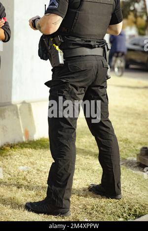 White male police officer cop standing on grass in a park during the day talking with person. Cop holding something in hand. Wearing bullet proof jack Stock Photo