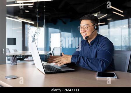 Portrait of a young male Asian teacher. He sits in the office at a table in a headset. He works with a laptop, teaches and explains to students via video call. Stock Photo