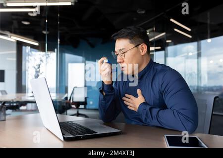 Young male Asian office worker having asthma attack at workplace, breathing hard. He sits at a table with a laptop, uses an inhaler. Stock Photo