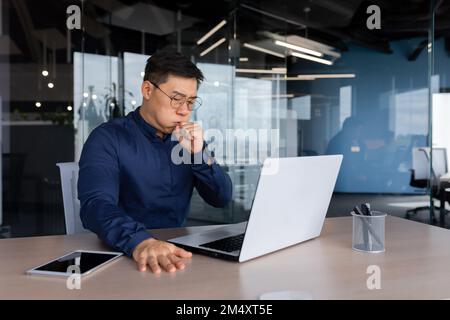 A young Asian businessman, lawyer fell ill at work. sits at the work table. coughs, covers his mouth with his hand, feels bad. Stock Photo