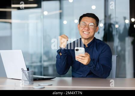 Portrait of a young happy businessman holding a phone in his hands, happy, showing a yes gesture. rejoices at winnings, online games, monetary rewards, good news. He looks at the camera smiling. Stock Photo