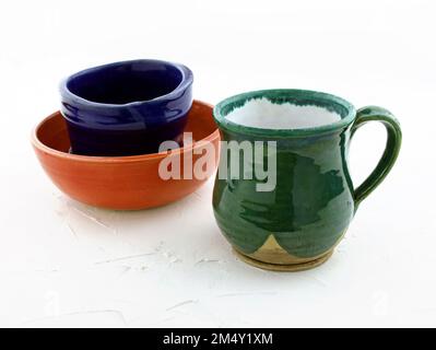 Color ceramic dishes on a white background. Cup made of green clay covered with glaze. Red ceramic bowl with black mug handmade. Empty cups in stack Stock Photo