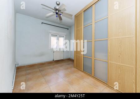 Empty room with a wall-to-wall built-in wardrobe with sliding doors made of oak wood and opaque glass, a fan with blades on the ceiling and light blue Stock Photo
