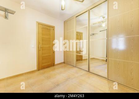 Empty room with wall-to-wall built-in wardrobe with oak sliding doors and mirrored ceiling fan, cream painted walls and light brown tiled floors Stock Photo