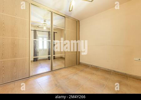 Empty room with wall-to-wall built-in wardrobe with oak sliding doors and mirrored ceiling fan, cream painted walls and light brown tiled floor Stock Photo