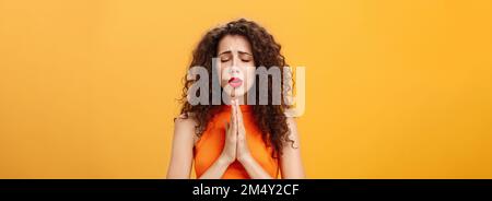 Nervous and concerned woman with curly hairstyle feeling hopeful praying with closed eyes and frowned eyebrows holding hands in pray near chest Stock Photo