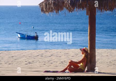 Young woman sitting on the beach, leaning against a man-made shade structure like a palm tree while glancing out at the water in a fisherman in a boat Stock Photo