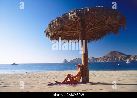 Younger woman. in a bathing suit and a hat sitting on the beach, leaning against a man-made shade device with a view of the ocean and rocky coastline Stock Photo