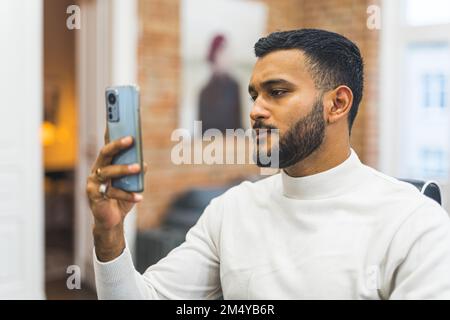 Well-groomed Indian man holding his phone on the eye level and sadly scrolling social media. Blurred background. Hairstylist salon interior. High quality photo Stock Photo