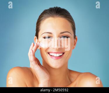 Because everybody loves glowing skin. Studio portrait of a beautiful young woman applying moisturizer to her face against a blue background. Stock Photo