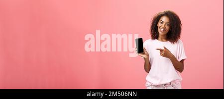 Suggest this model of phone. Happy charming african american curly-haired woman in stylish outfit holding smarpthone and pointing at device screen Stock Photo