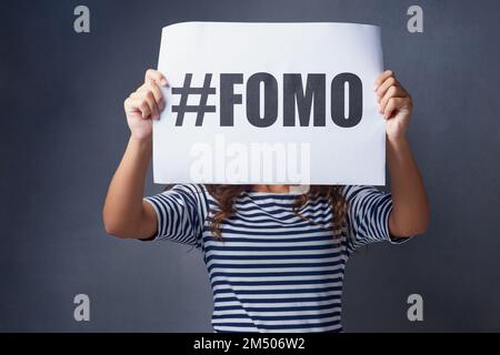 I feel so excluded. Studio shot of a young woman holding a sign with FOMO printed on it against a gray background. Stock Photo