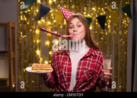 Smiling young woman in birthday cap blowing party horn at home, holding champagne and cake. Positive lady celebrating holiday at home atmosphere. Fest Stock Photo