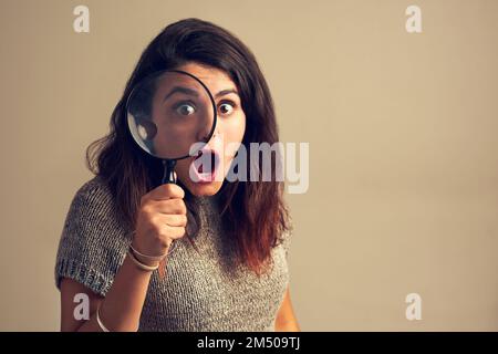 When you finally find what youve been stalking for. Studio portrait of a young woman looking shocked while using a magnifying glass against a brown Stock Photo