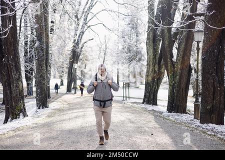 Sporty father carrying his infant son wearing winter jumpsuit and cap in backpack carrier walking in city park in winter. Stock Photo