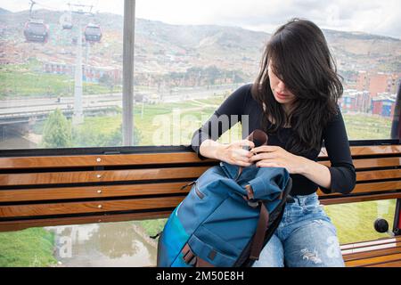 A young woman, wearing a black shirt and jeans, sitting on a bench and holding her backpack, waiting for a gondola ride, the concept of travelling Stock Photo