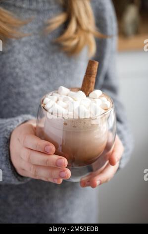 a girl in a gray sweater holds a glass of cocoa, marshmallows and cinnamon stick  in the early morning against the background of the kitchen. Stock Photo