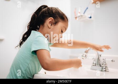 A cute preschool girl practicing good hygiene using the bathroom sink as she washes up Stock Photo