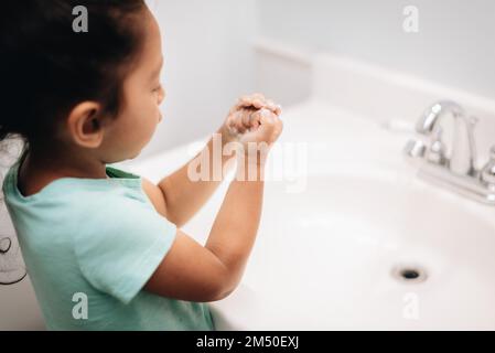 A cute preschool girl practicing good hygiene using the bathroom sink as she washes up Stock Photo