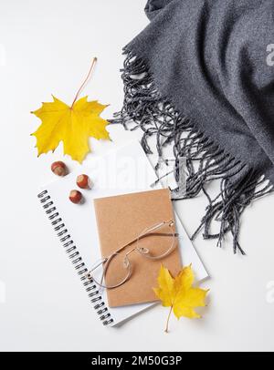 Concept cozy autumn flat lay with yellow maple leaves on notepad with eyeglasses and gray scarf  on a white table. Top view and copy space. Stock Photo