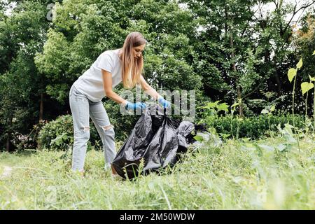 Young caucasian woman cleaning-up public park or forest of plastic garbage. Volunteer picking up plastic bottle in woods. Green and clean nature Stock Photo