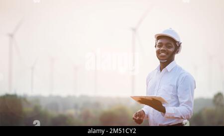 African engineer windmills wearing white hard hat and using tablet with wind turbine on sunset Stock Photo