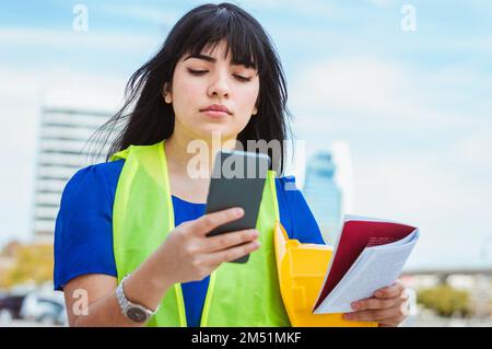 Young Caucasian Venezuelan woman, engineering worker, standing on the street, with her phone in her hand attentively reading a message from her boss, Stock Photo