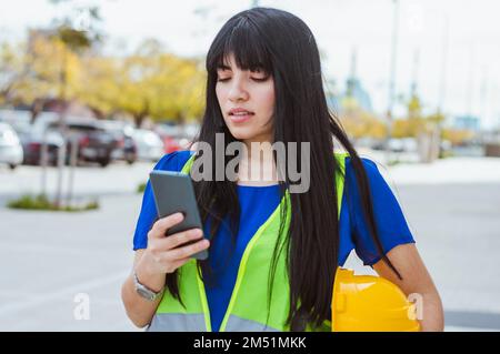 young engineer woman standing on the street resting, holding the protection helmet in her hands and using her phone, watching the mobile and typing, s Stock Photo